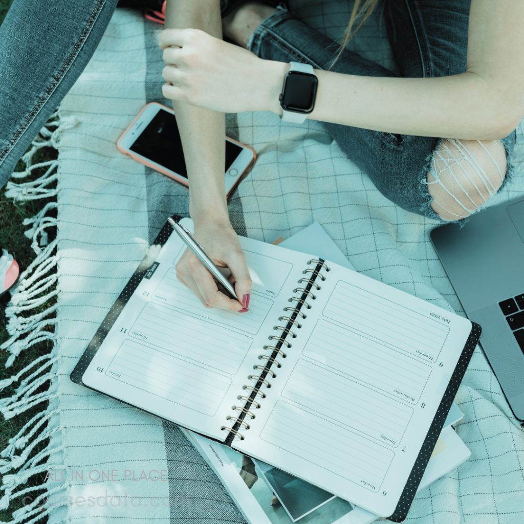 Writing in a planner. Wearing a smartwatch. Smartphone beside the planner. Sitting on a picnic blanket. Laptop partially visible nearby.