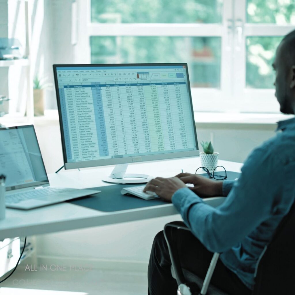 Person working on spreadsheet. Two computer monitors visible. Bright, airy office environment. Green plant in small pot. Desk with minimalist design.