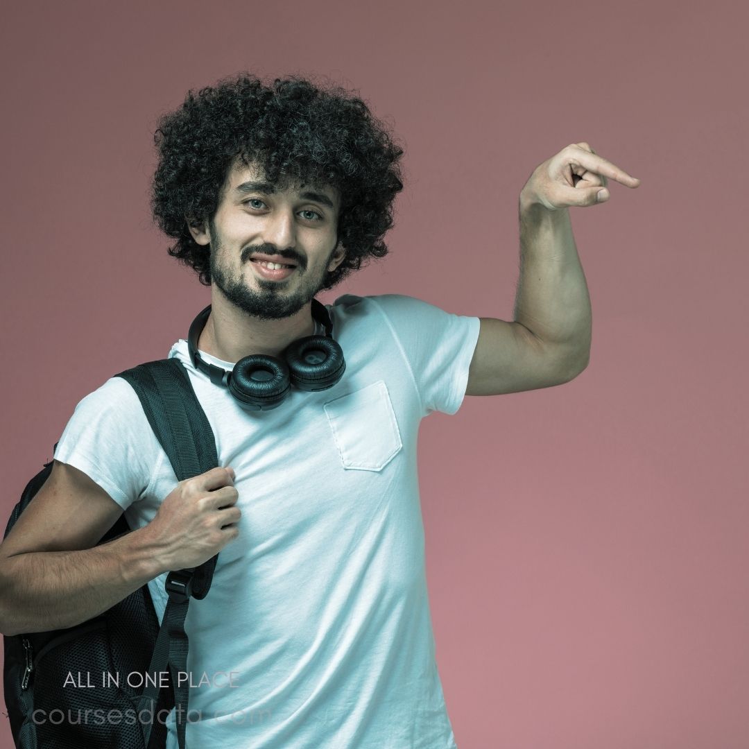 Smiling man with curly hair. Wearing white t-shirt. Backpack slung over shoulder. Headphones around his neck. Pointing with one hand.