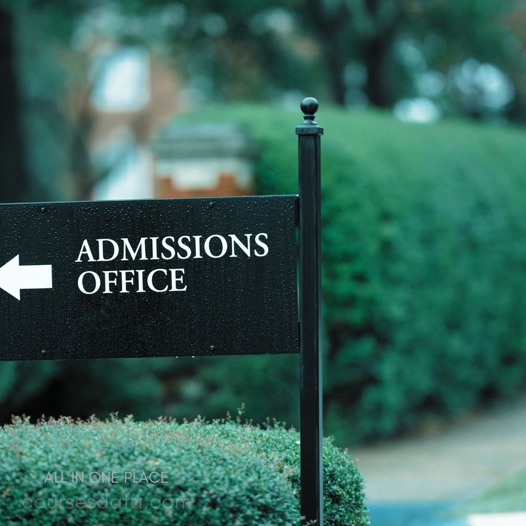 Black sign, white text. "Admissions Office" displayed. Arrow pointing left. Surrounded by green bushes. Background slightly blurred.