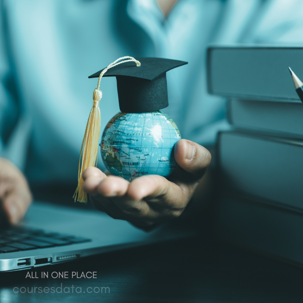 Graduation cap on globe. Person holding globe. Laptop and books nearby. Blue shirt in background. Focus on education theme.