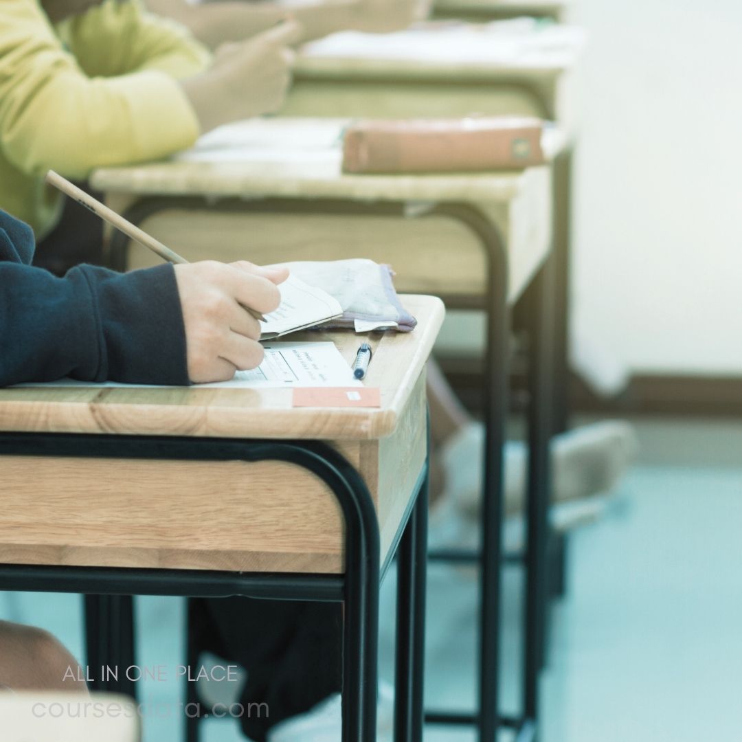 Student writing on desk. Classroom setting, multiple desks. Pencil and stationery visible. Casual attire, focused expression. Soft, natural lighting ambiance.