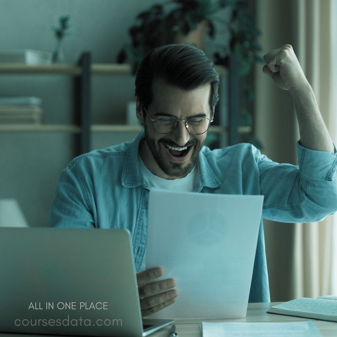 Man celebrating with paper. Joyful expression, raised fist. Laptop and notebook beside him. Bright indoor setting, greenery visible. "All in one place" text.