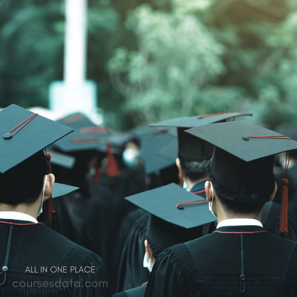 Graduates wearing black caps. Ceremony in outdoor setting. Some masks visible on faces. Green background with blurred trees. Focus on backs of graduates.