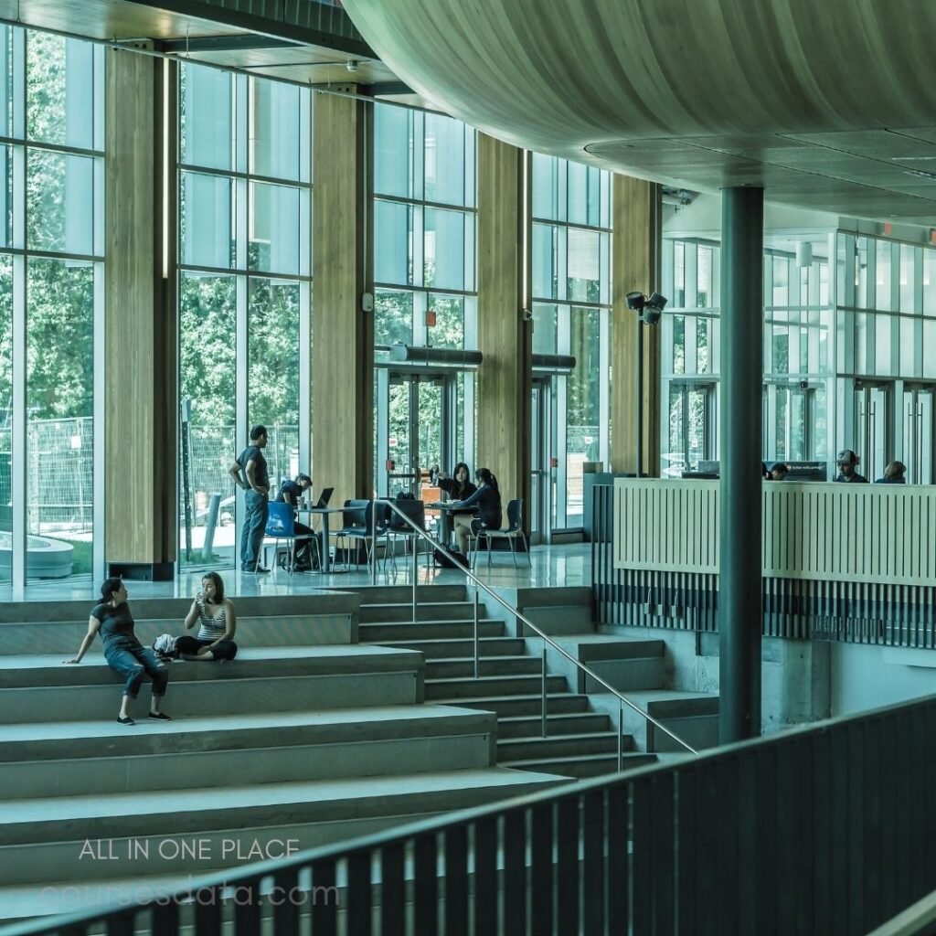 Modern interior with large windows. People sitting on steps. Group collaborating at tables. Natural light enhances atmosphere. Wood accents throughout design.