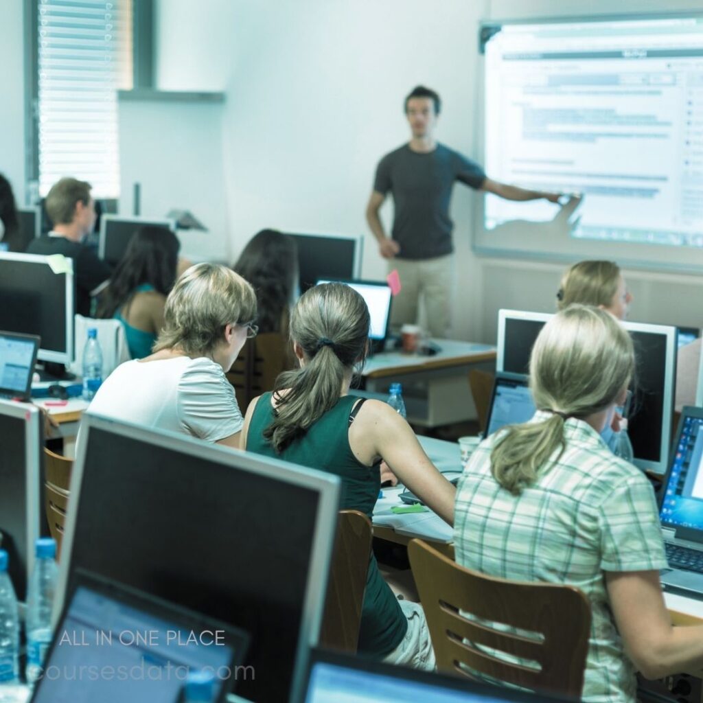 Classroom with students studying. Instructor presenting on screen. Students engaged with computers. Bright, modern learning environment. Diverse group of learners.