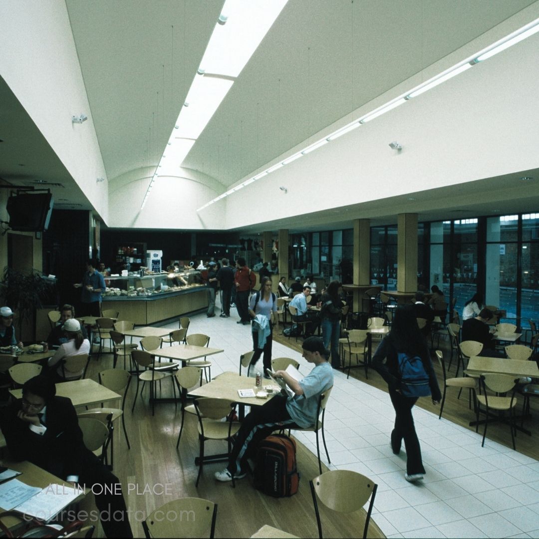 Cafeteria filled with students. Bright, open space design. Tables surrounded by chairs. Counters with food options. Natural light from large windows.
