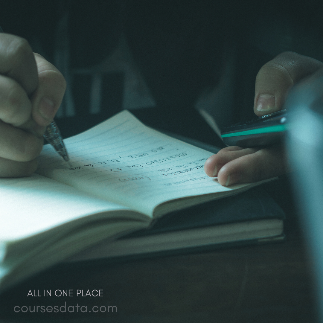Hand writing in notebook. Dark background environment. Additional pen and device nearby. Focused study or note-taking activity. Notebook pages visible with text.