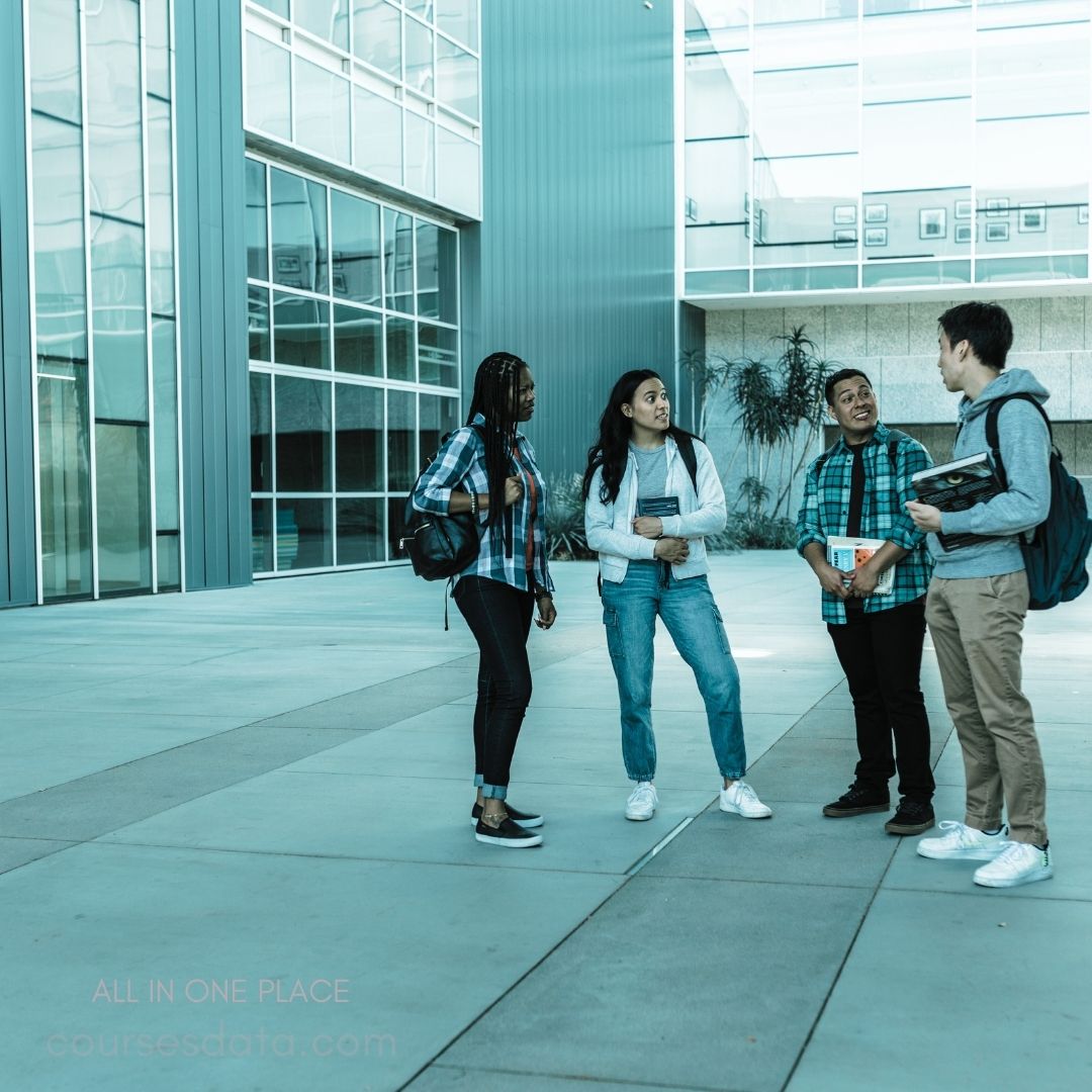 Four students chatting outdoors. Modern building in background. Casual attire with backpacks. Diverse group of friends. Engaged and smiling expressions.