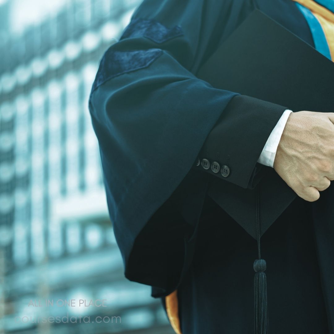 Graduate in academic regalia. Holding diploma folder tightly. Urban background, blurred buildings. Blue and gold robe details. Formal shirt and cufflinks visible.