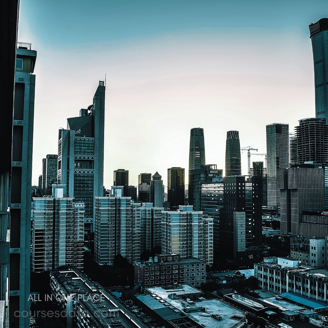 City skyline at dusk. Modern high-rise buildings. Muted blue and green tones. Urban landscape with cranes. City lights starting to glow.