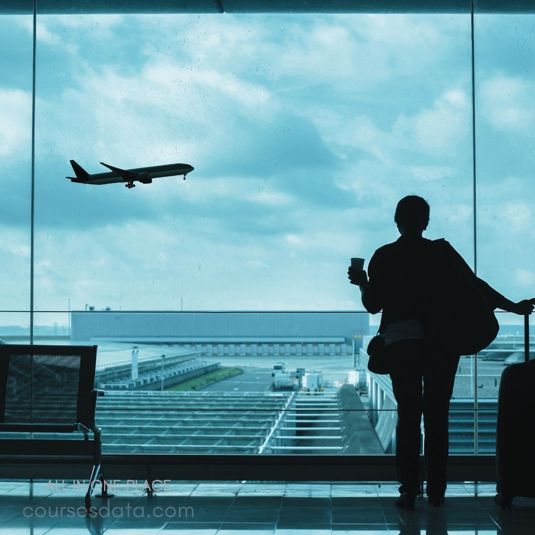 Person waiting at airport window. Airplane flying outside. Suitcase and coffee cup nearby. Cloudy sky reflects through glass. Empty terminal seats visible.