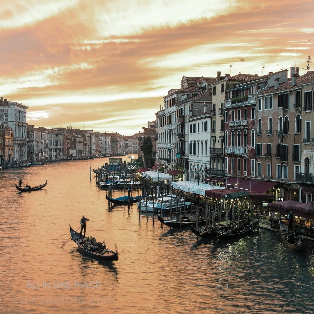 Venetian canal at sunset. Gondolas on tranquil waters. Historic buildings lining the shore. Warm, colorful sky reflections. Outdoor dining along the canal.