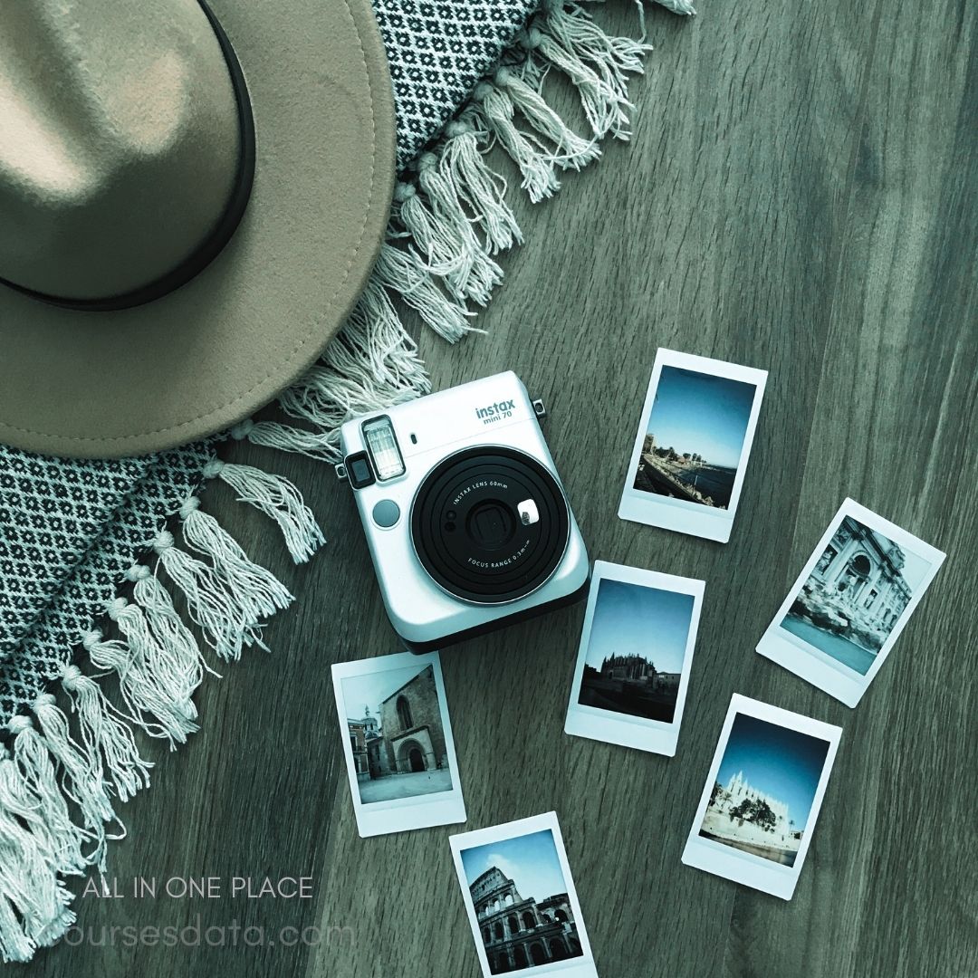 Instant camera and photos. Tan wide-brimmed hat. Textured fabric background. Polaroid photos scattered around. Wooden surface beneath items.