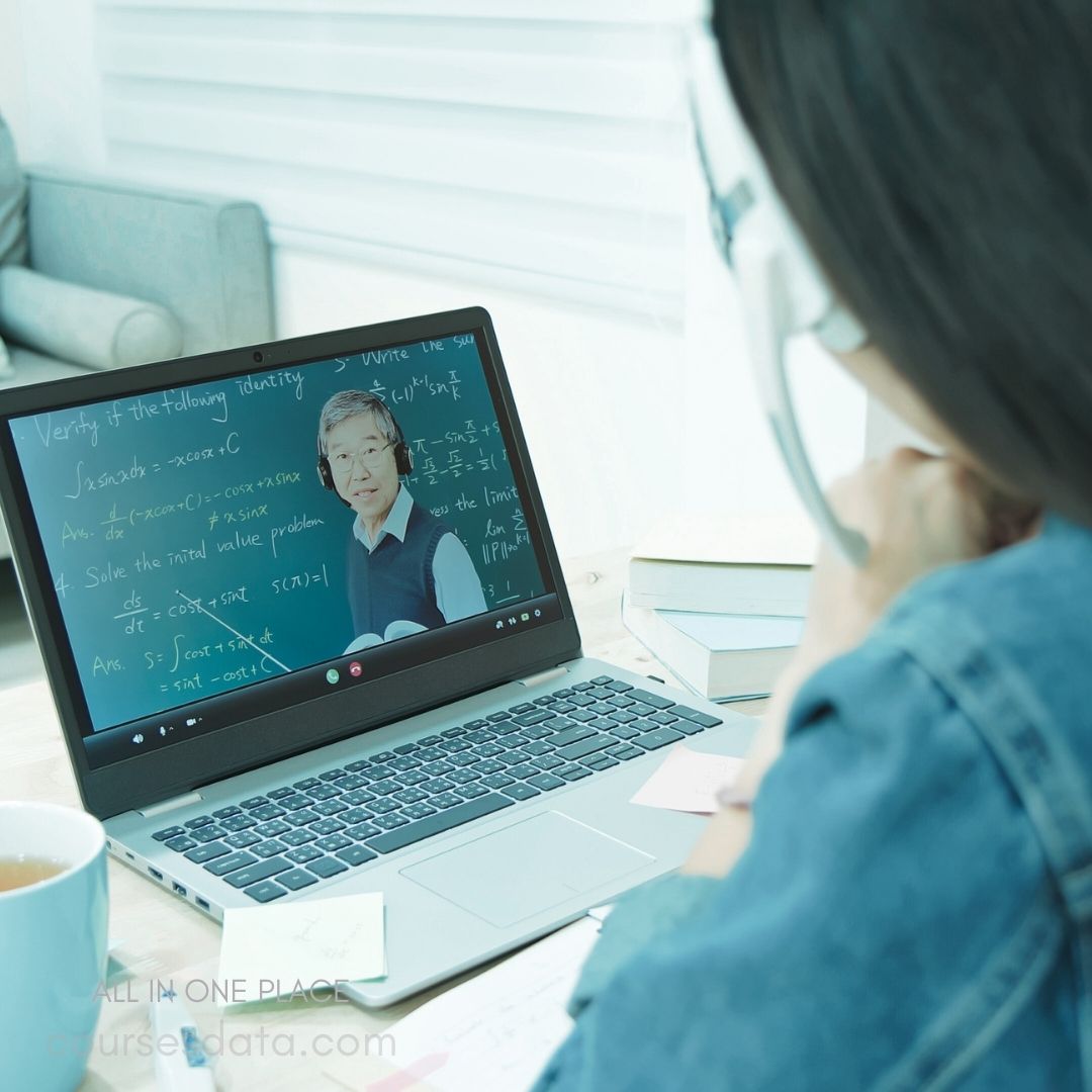 Student watching online lecture. Teacher explaining math concepts. Mathematical equations on screen. Cup of tea on desk. Books and notes nearby.