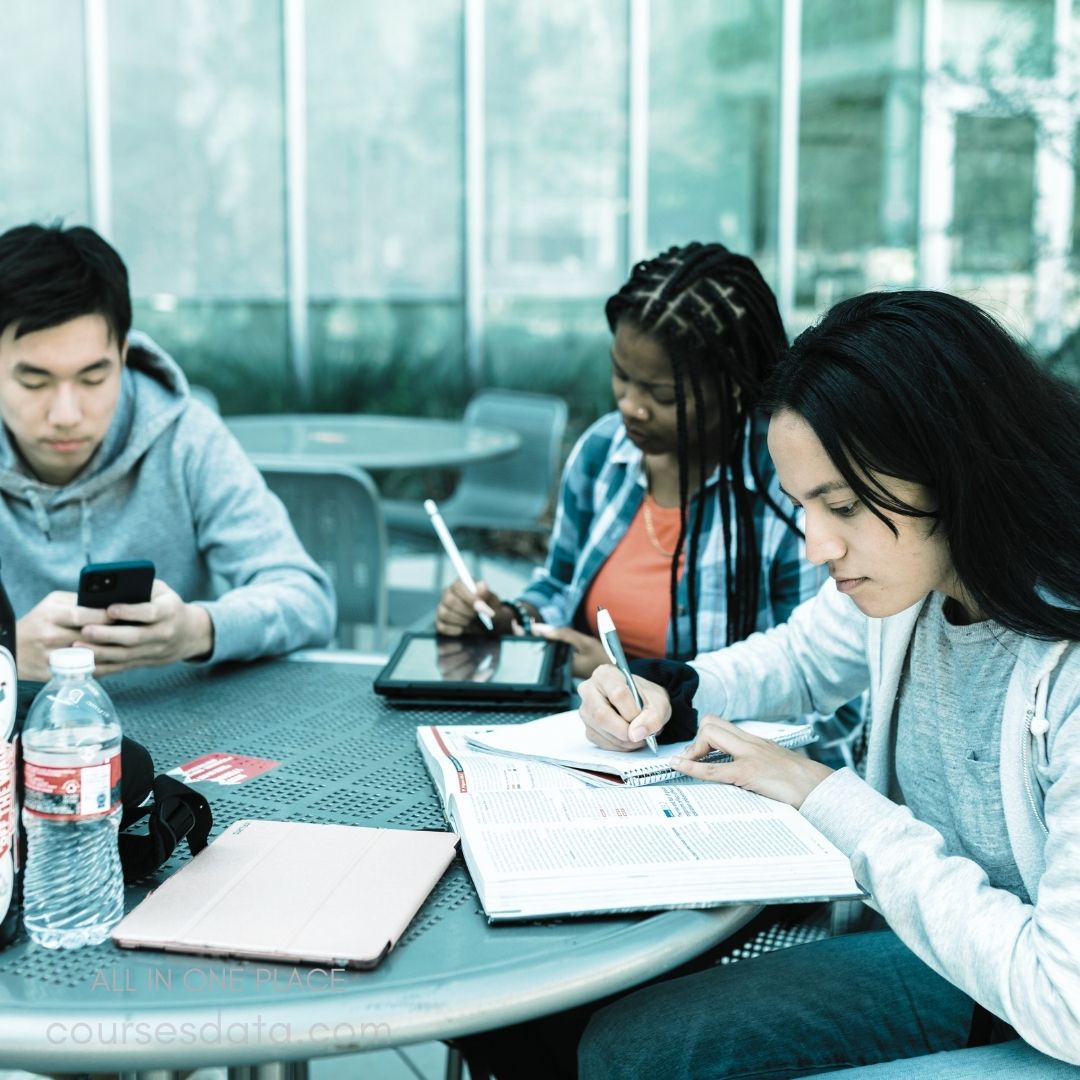 Group studying outdoors. Person on phone. Individual using tablet. Student taking notes. Textbook open on table.