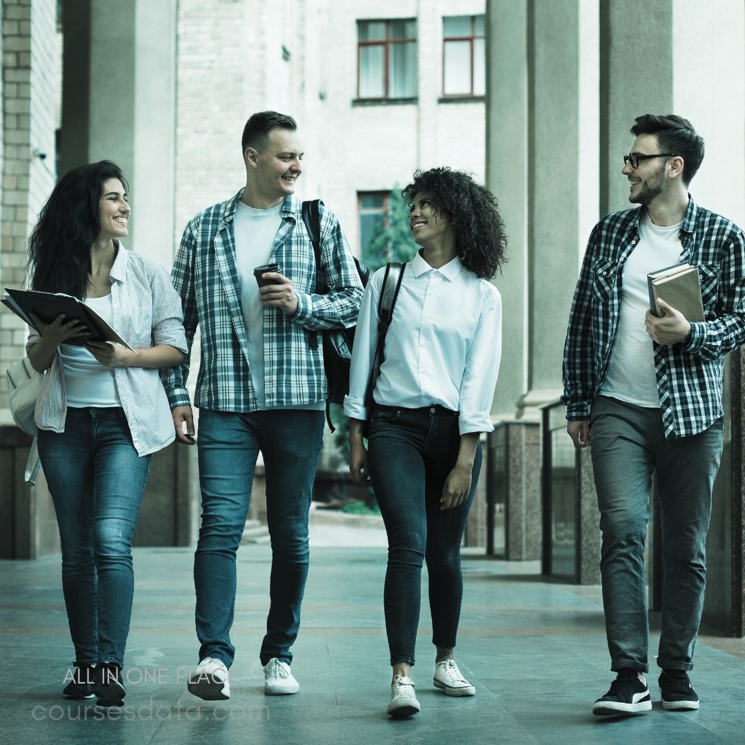 Four smiling friends walking. Casual outfits with backpacks. Bright outdoor hallway setting. Engaged in friendly conversation. Carrying notebooks and drinks.