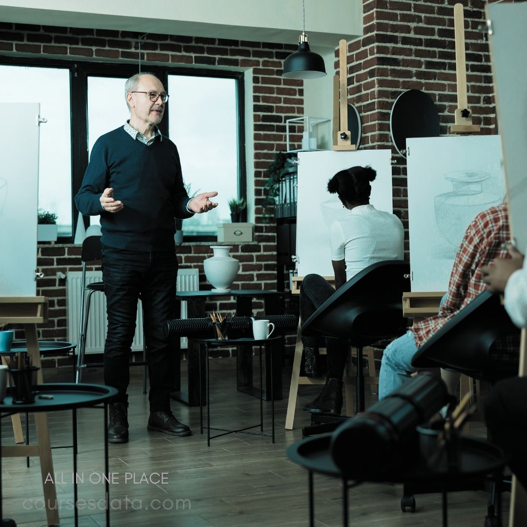 Art class in a studio. Instructor speaks to students. Easels display sketch drawings. Brick wall and large windows. Various art supplies on tables.