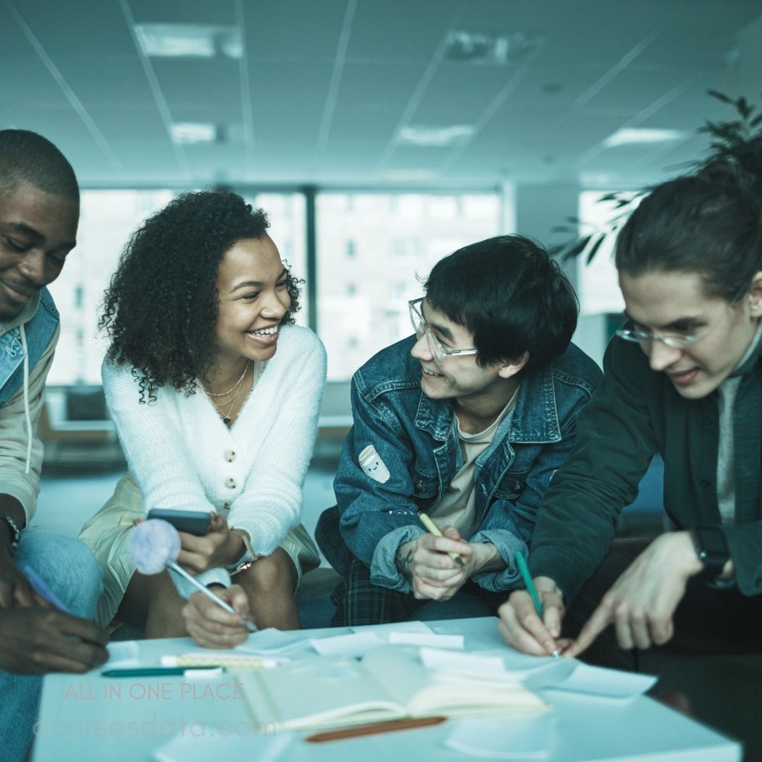 A diverse group of four young adults is engaged in a collaborative discussion around a table scattered with notes and pens. They appear to be brainstorming or working on a project, with smiles and laughter indicating a friendly atmosphere. The setting is a bright, modern office space with large windows in the background.