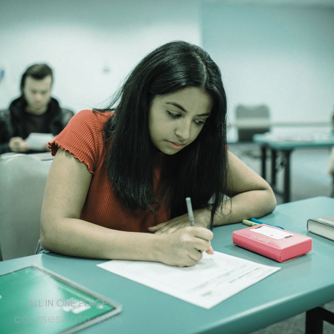 A student with long, dark hair is sitting at a green table, focused on writing on a sheet of paper. She is wearing a red top and is surrounded by a classroom setting, with a classmate visible in the background. A pink box and some pens are placed on the table next to her.
