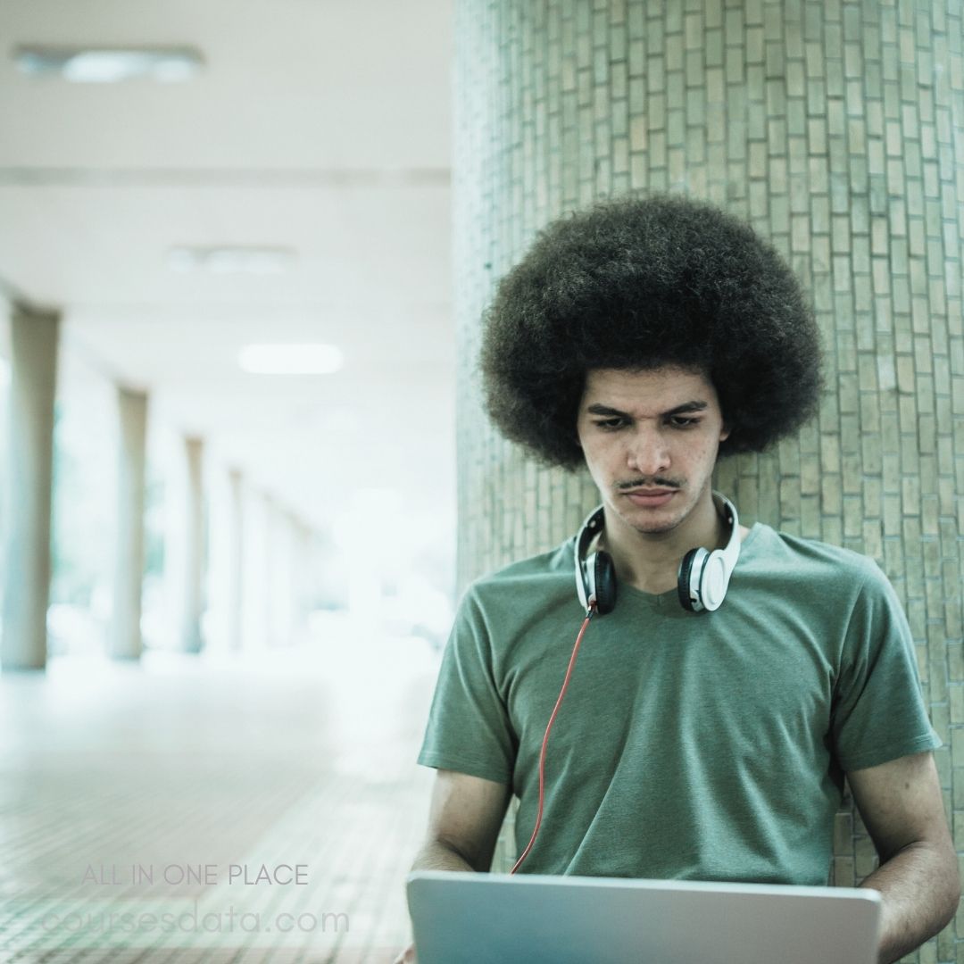 A young man with a large, textured afro sits against a textured column, focused on his laptop. He wears a plain green t-shirt and has white headphones around his neck, with a red cord leading to the device. The setting is a spacious, well-lit corridor, suggesting a public or educational environment.