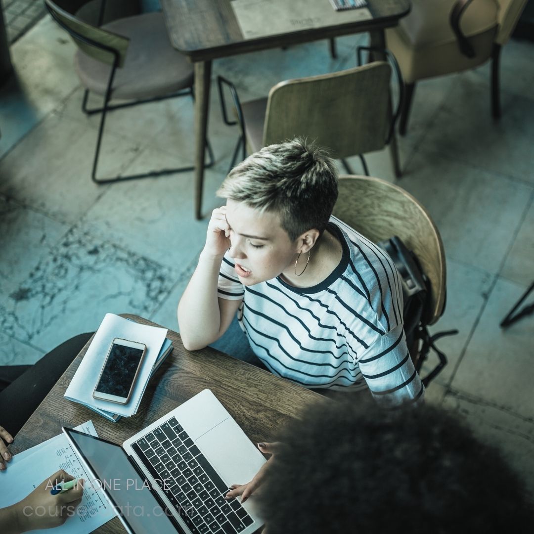 A young woman with short, light hair sits at a wooden table in a cafe, looking focused as she types on a laptop. She wears a striped shirt and rests her chin on her hand, appearing deep in thought. A smartphone and some notebooks are placed nearby, along with a partially visible hand from another person, suggesting collaboration or discussion. The cafe features a cozy atmosphere with wooden furniture and warm lighting.