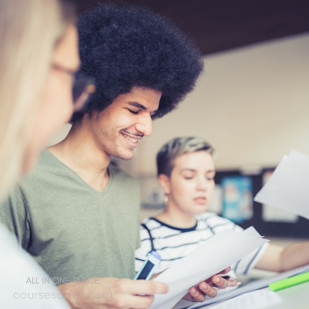 A young man with an afro smiles while reviewing papers in a collaborative setting. He is wearing a green shirt and is engaged in a discussion with two others: a woman with blonde, wavy hair on the left, partially visible, and another woman with short, textured hair in a striped shirt on the right. Papers and highlighters are scattered on the table, reflecting a study or work session.