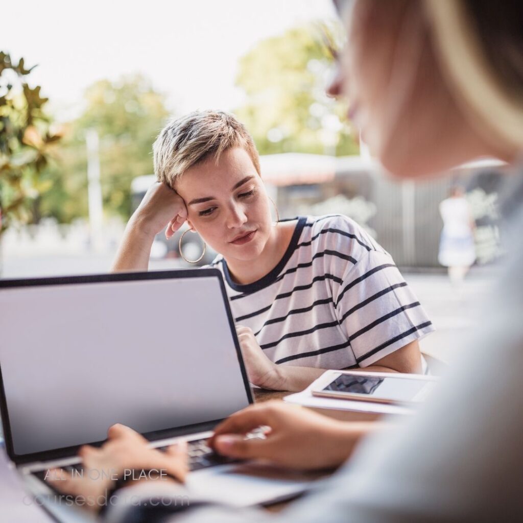 Two young women are seated at an outdoor table, engaged in a focused discussion. One woman, wearing a striped t-shirt and large hoop earrings, leans forward with her chin resting on her hand, concentrating on the laptop in front of her. The second woman, partially visible, types on a laptop and appears to be sharing information. The background features greenery and a hint of urban scenery. The overall atmosphere is casual and collaborative.