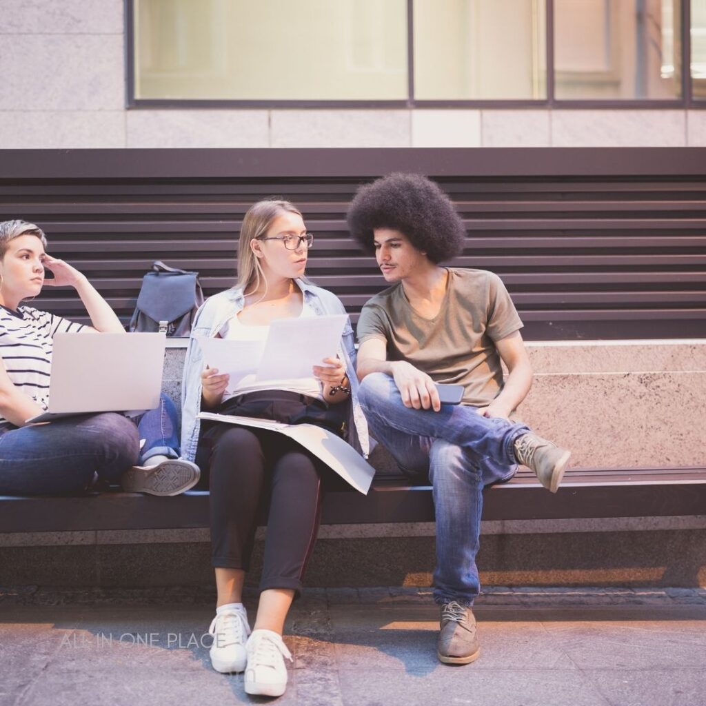 Three young adults are seated on a bench in an urban setting. One woman, wearing a striped shirt and looking pensive, is working on a laptop. The woman in the middle, with long straight hair and glasses, holds documents, engaged in conversation with a man with an afro hairstyle, who is casually dressed in a grey t-shirt and jeans, holding a phone. They appear to be collaborating on a project. In the background, there's a backpack resting against the bench.