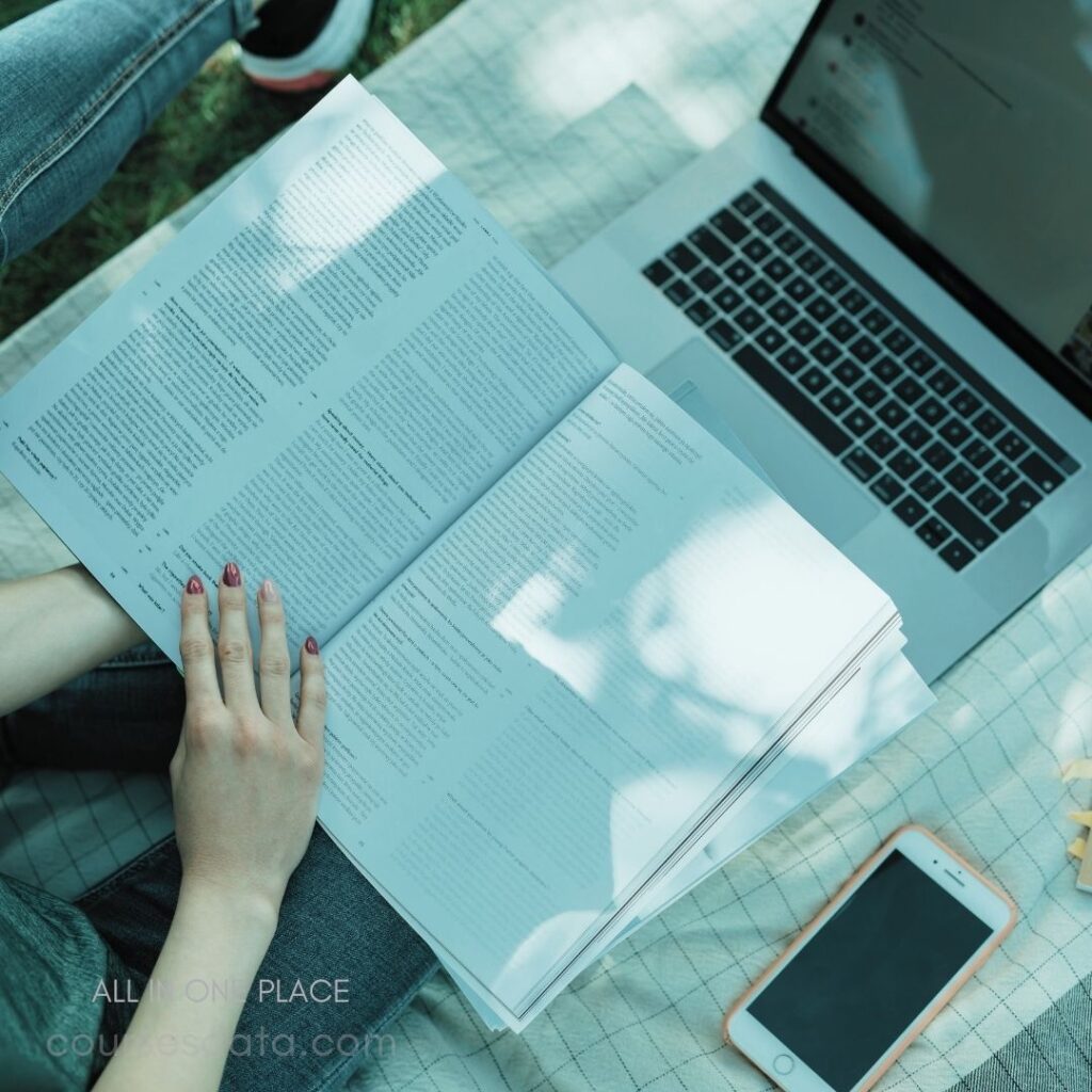 Person reading open book. Laptop beside on blanket. Smartphone placed on ground. Sunlight casting gentle shadows. Casual outdoor study session.