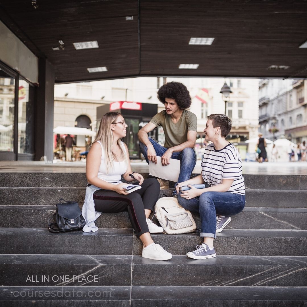 Three young adults are sitting on a set of stone steps in a bustling urban area. The woman on the left has long blonde hair and wears a white tank top and black leggings, holding a notebook. The man in the middle has curly hair and is dressed in a green shirt and jeans, looking at the others. The woman on the right has short hair and is wearing a striped t-shirt and jeans, holding a cup. They seem to be engaged in a friendly discussion, with shops and people in