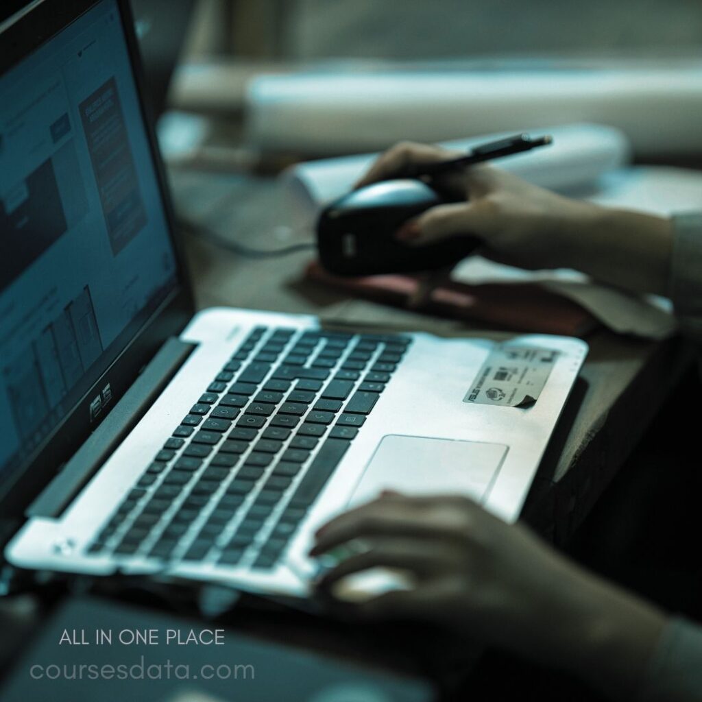 Person using laptop and mouse. Dimly lit workspace, focused task. Hand holding pen near mouse. Screen displays digital content. Desk cluttered with papers.