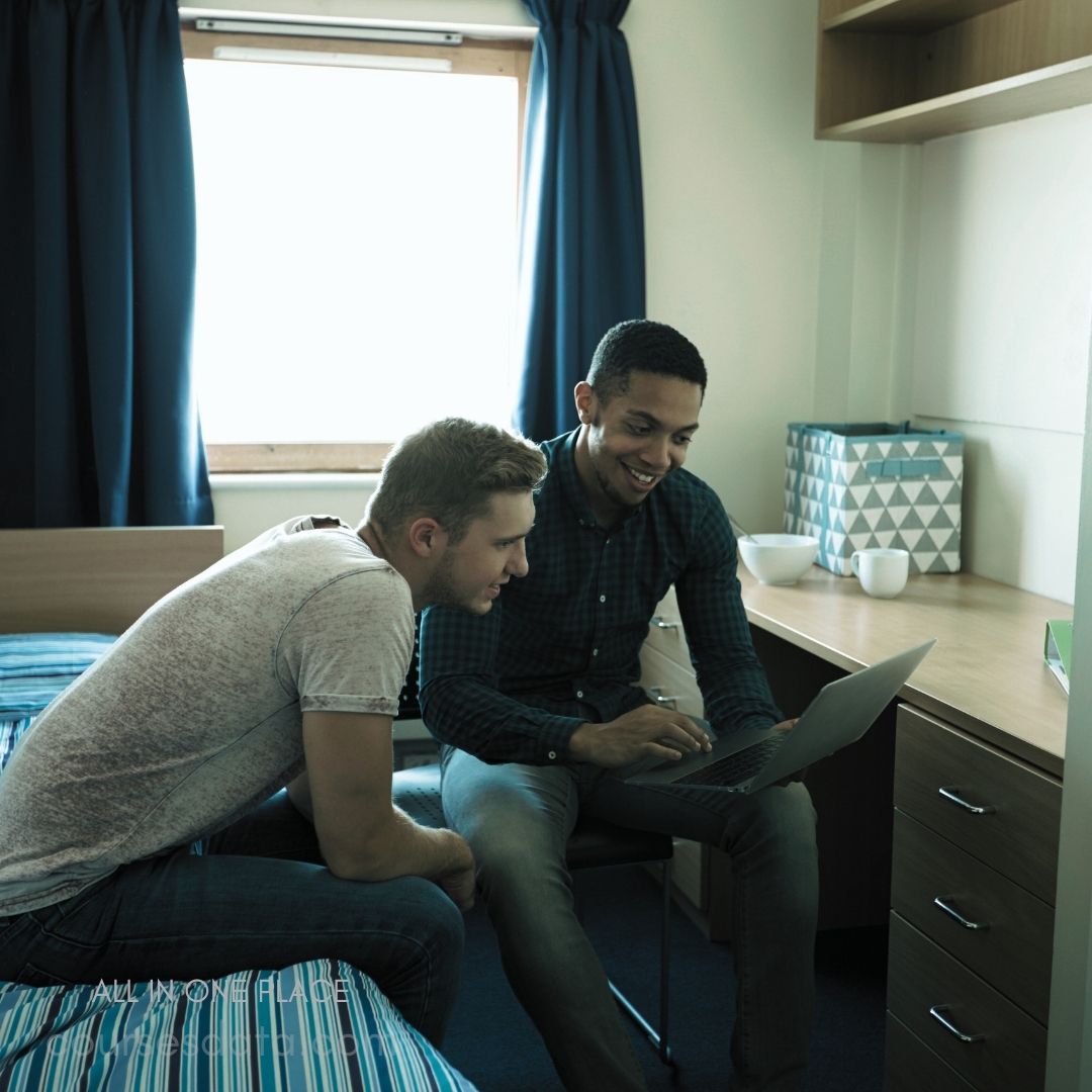 Two friends sharing laptop. Bright room with blue curtains. Wooden furniture and striped bedding. Smiling, looking at screen. Casual attire, relaxed atmosphere.