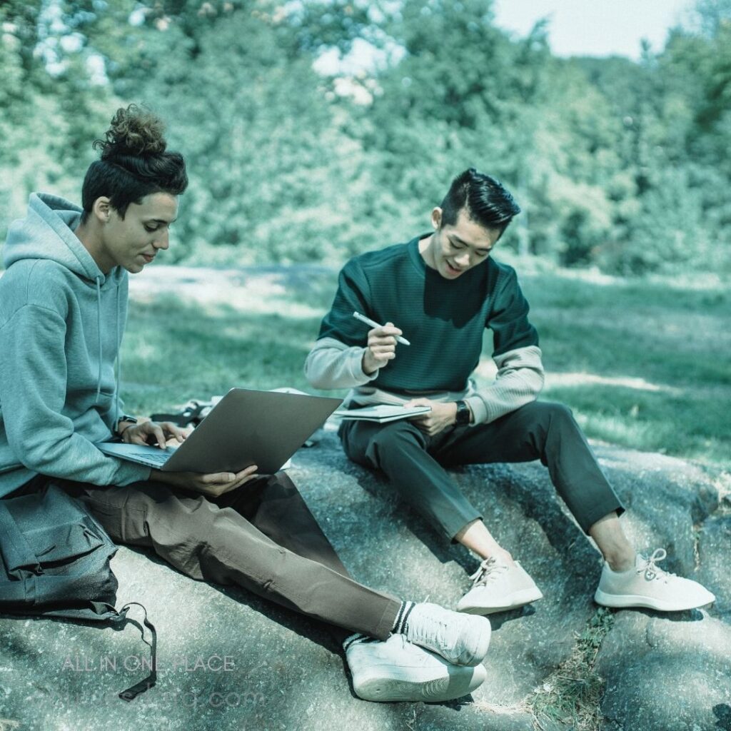 Two young men studying outdoors. One using a laptop, the other sketching. Surrounded by green trees. Sunlight filtering through branches. Casual clothing and relaxed atmosphere.