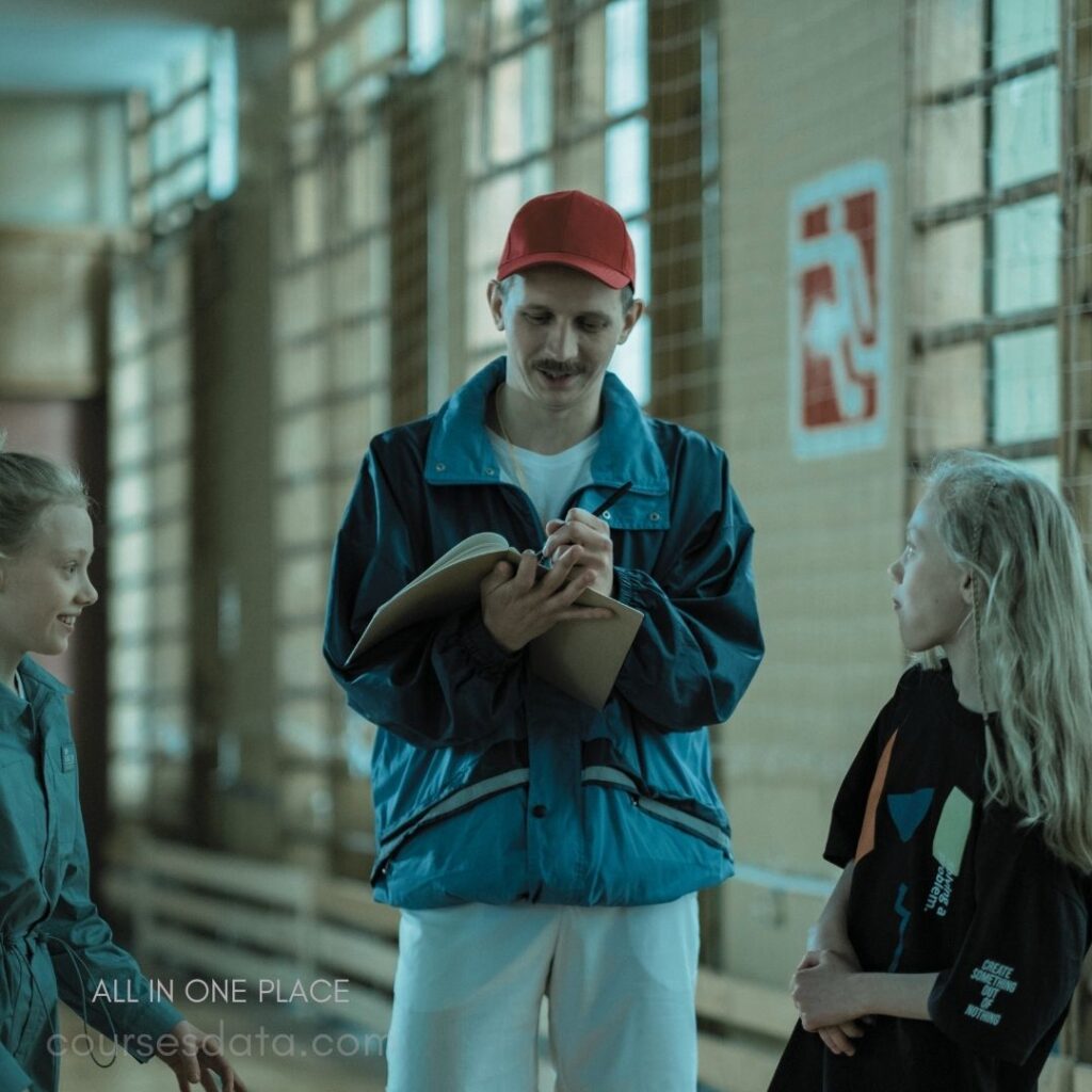 Man in blue jacket talking. Two girls listening attentively. Indoor setting with windows. Notebook and pen in hand. Red cap worn by man.