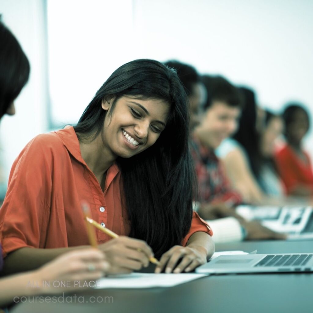 Smiling woman writing notes. Casual classroom setting. Students focused on tasks. Laptop and notebooks visible. Bright, airy environment.