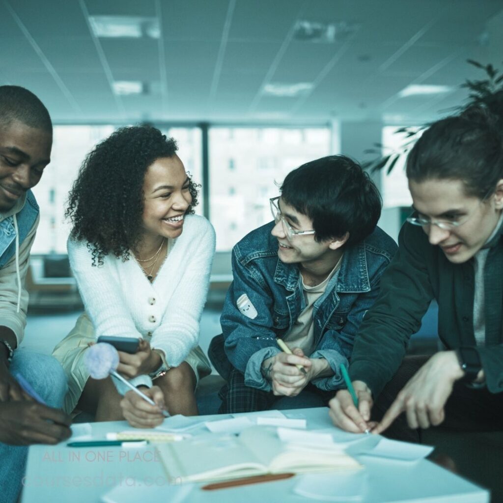 Four friends collaborating indoors. Smiling and engaging discussion. Notebooks and paper scattered. Bright, modern office setting. Casual attire, diverse group.