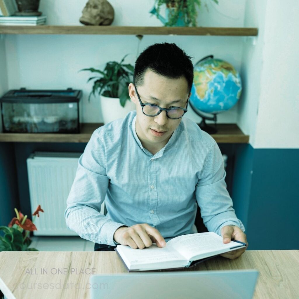 Man reading a book. Blue glasses and shirt. Indoor study environment. Potted plants in background. Globe and skull decor.