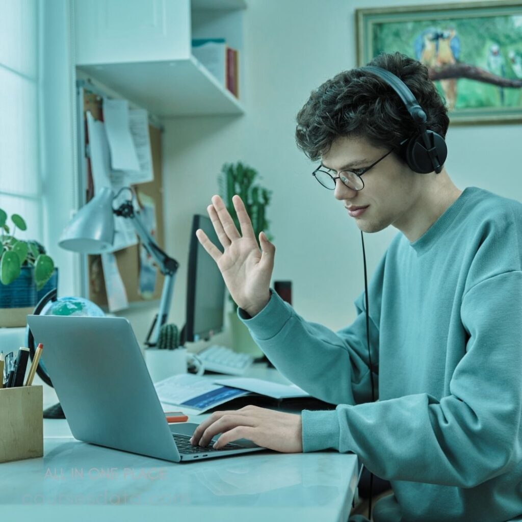 Young man gestures while studying. Wearing headphones and glasses. Laptop open on desk. Plant and notes in background. Bright, soft color palette.