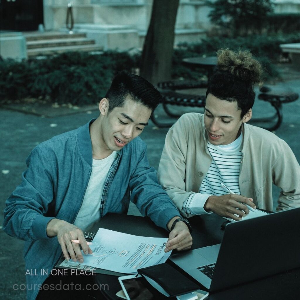 Two young men collaborating. Studying documents together. Laptop and notes on table. Outdoor setting with greenery. Engaged in focused discussion.