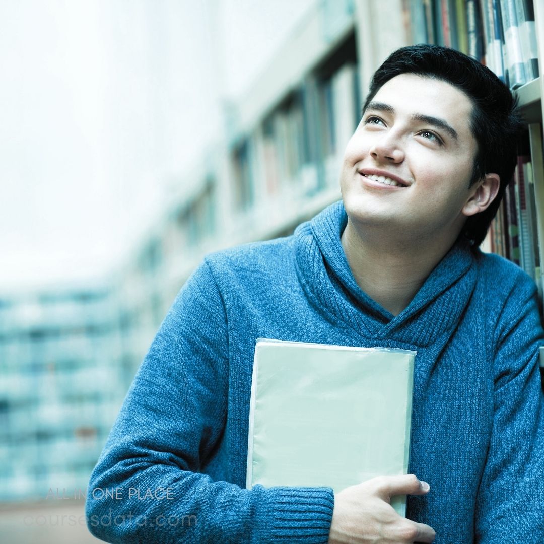Smiling young man in library. Holding a book tightly. Wearing a blue sweater. Looking upward thoughtfully. Bookshelves in background blurred.