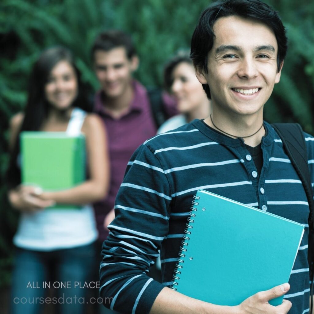 Smiling student holding notebook. Friends blurred in background. Green foliage backdrop. Casual attire with stripes. Campus setting atmosphere.