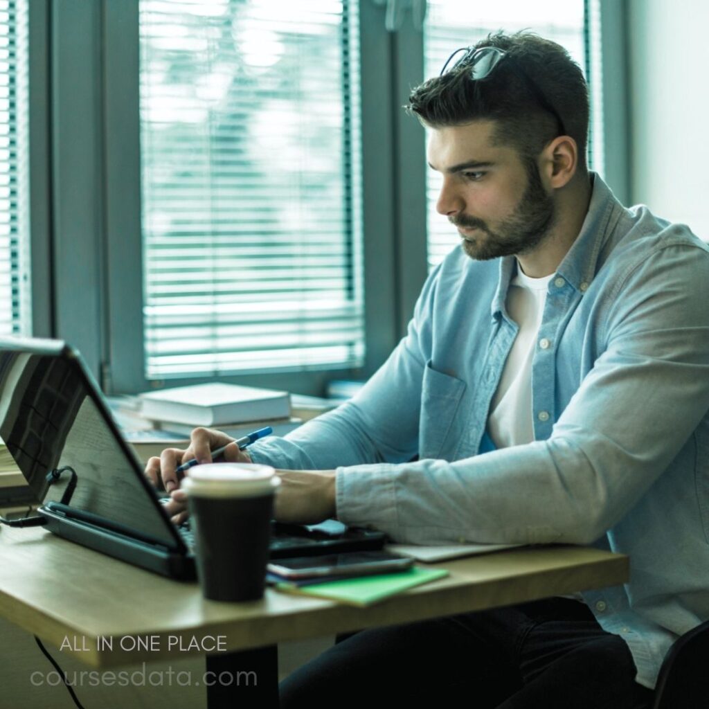 Young man working intently. Sitting at a wooden desk. Using a laptop and pen. Coffee cup beside him. Sunlight through the window.