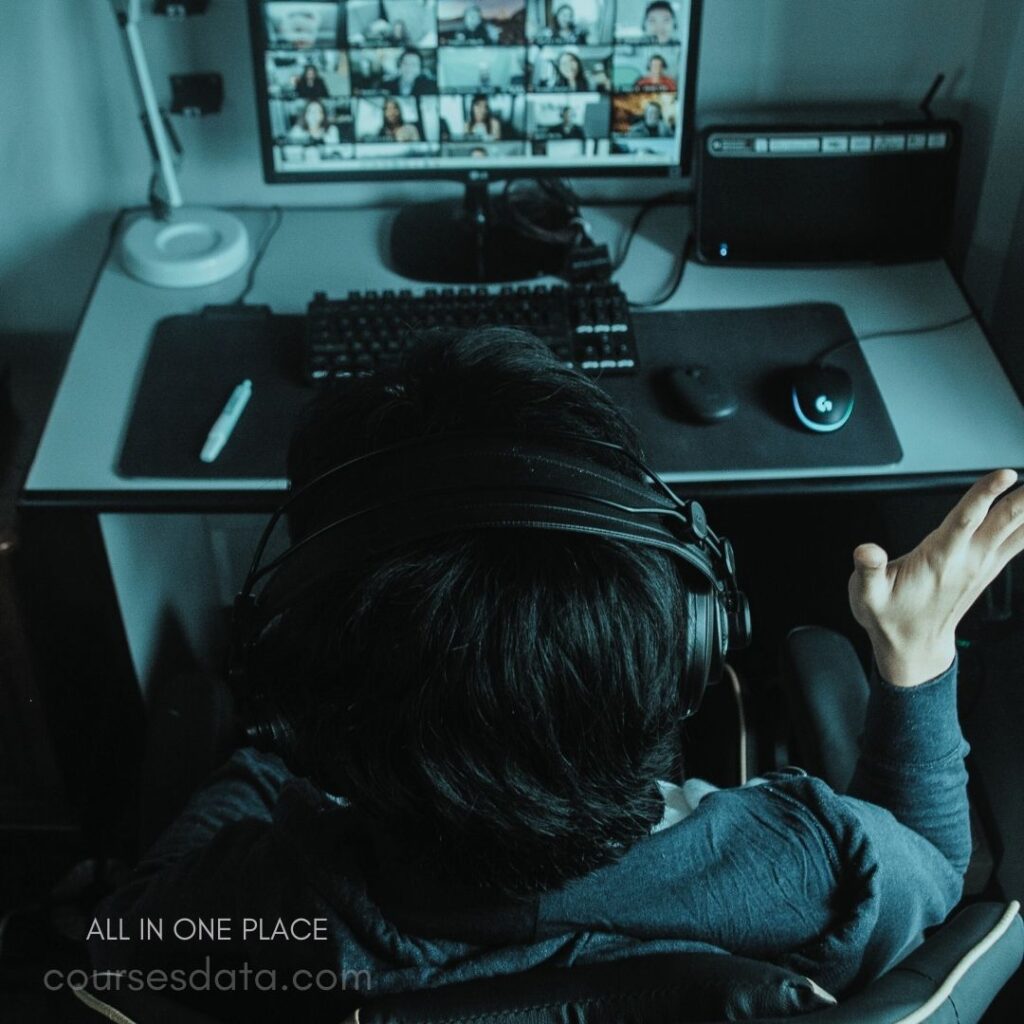 Person wearing headphones gesturing. Computer screen with video calls. Desk with keyboard and mouse. Dark, focused workspace environment. Engaged in virtual meeting.