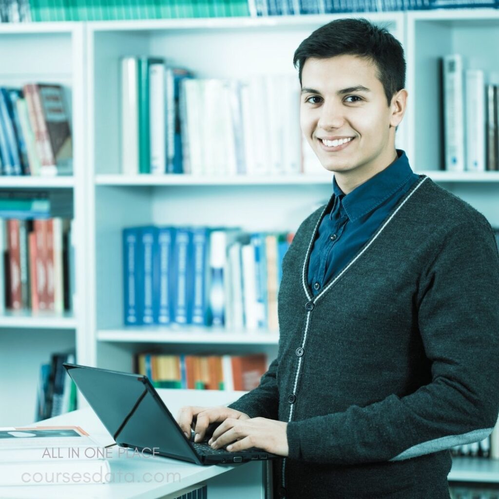 Smiling young man at laptop. Bookshelves filled with books. Casual outfit with cardigan. Modern, well-lit study environment. Engaged in productive work.