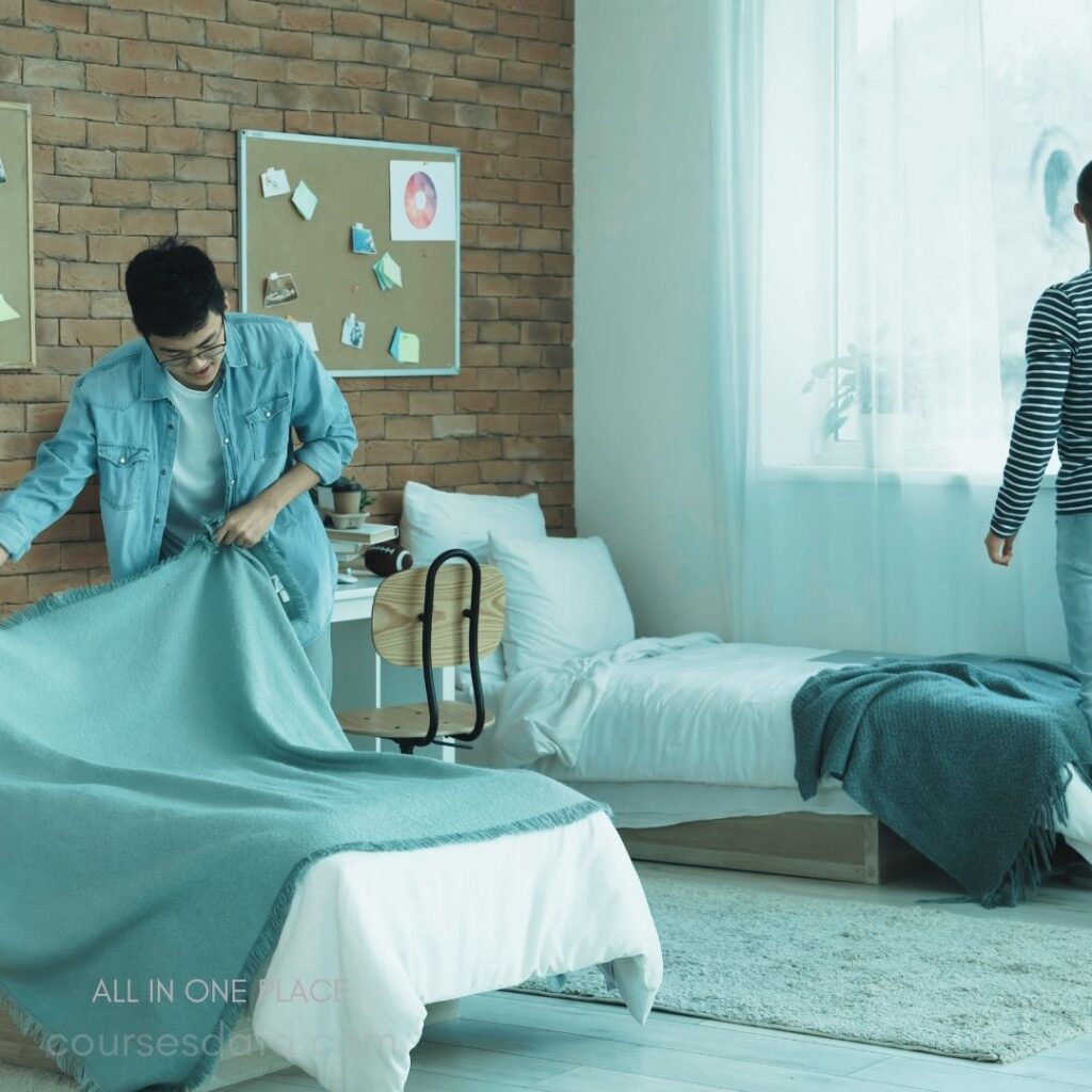 Two people arranging bedding. Cozy bedroom with brick wall. Light blue blanket on bed. One person smiles while working. Bright, airy natural light.