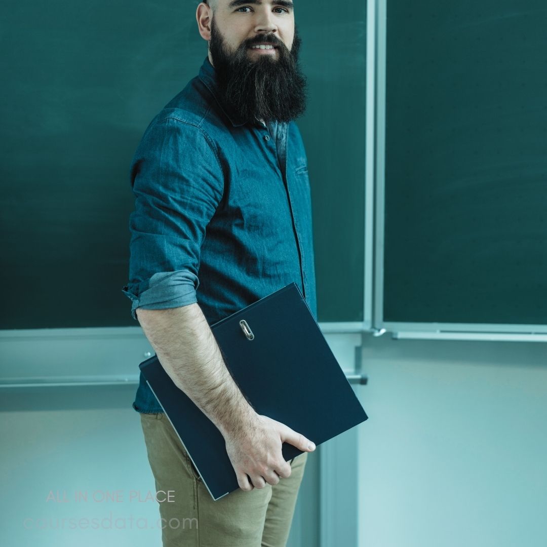 Bearded man holding clipboard. Wearing a blue shirt. Standing in a classroom. Green chalkboard in background. Casual professional appearance.