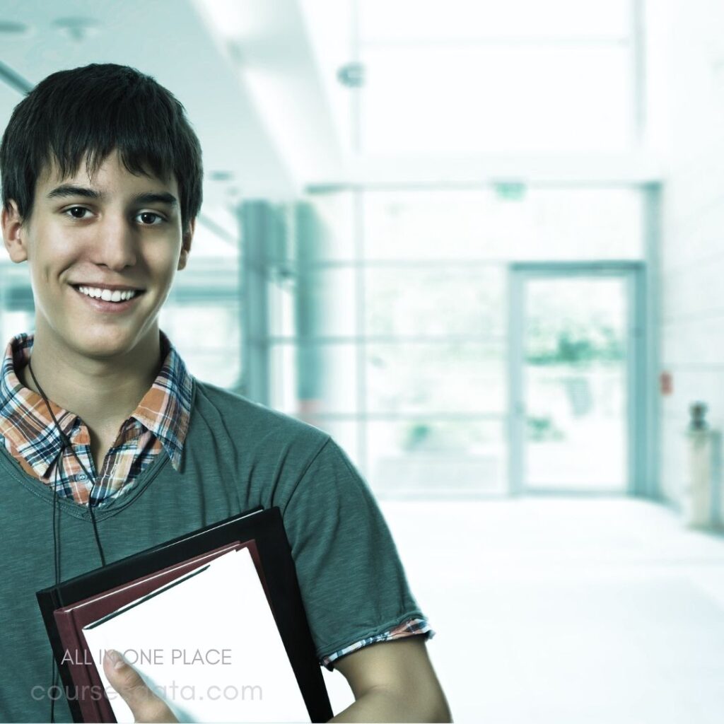 Smiling young man, holding folders. Bright, modern hallway background. Casual attire, plaid shirt visible.