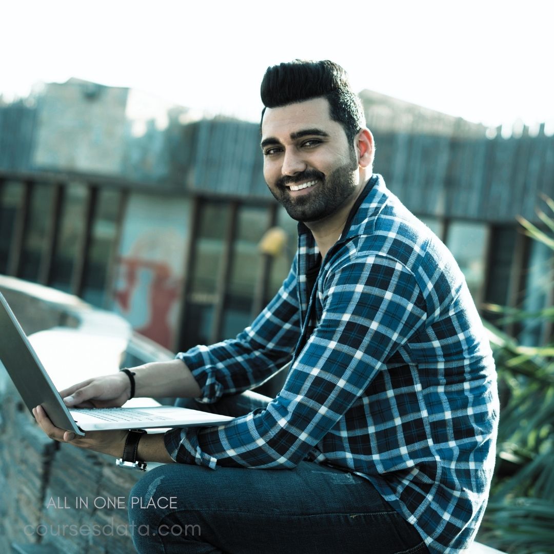 Smiling man with laptop. Wearing blue plaid shirt. Outdoor setting with greenery. Casual, relaxed pose. Bright, sunny background.