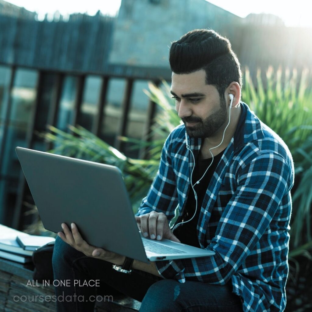 Man using laptop outdoors. Wearing headphones, focused expression. Surrounded by greenery, bright atmosphere. Casual plaid shirt, relaxed posture. Sitting on wooden surface.